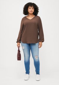 Une femme aux cheveux bouclés porte un haut marron à manches longues et un jean bleu déchiré, tenant un sac à main bordeaux, se tenant contre un fond blanc.
