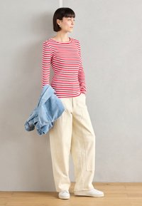 Red and white striped long-sleeve shirt paired with loose cream-colored trousers. Holding a folded light blue denim jacket. White sneakers.