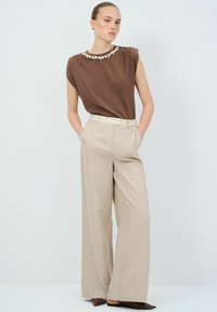 Bruine mouwloze top met een gestructureerde halslijn, gecombineerd met hoog getailleerde beige wijde broek, voorzien van een riem en een subtiele gemêleerde textuur.