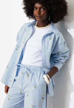 Femme aux grands cheveux bouclés portant une chemise rayée bleu clair et un pantalon à petits motifs floraux ainsi qu'un haut blanc, debout avec la main dans la poche.