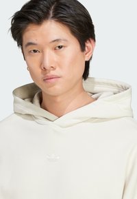 Hoodie color crema con coulisse e dettagli a coste. Presenta un logo discreto al centro. Tessuto morbido e liscio.