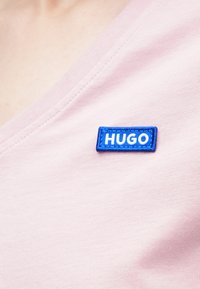 Rózsaszín rövid ujjú ing mély V-nyakkivágással, a mellrészen kék téglalap alakú folttal, melyen fehér betűkkel "HUGO" felirat látható, rávarrva.