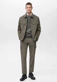 Olivengrüne Cargo-Jacke mit zwei Brusttaschen, kombiniert mit passenden Slim-Fit-Hosen und schwarzen Sneakers. Glattes Material mit minimaler Textur.