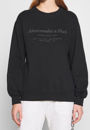 Sweat-shirt noir Abercrombie & Fitch avec le nom de la marque et un texte brodés, porté par une personne en pantalon gris clair avec des détails en patch.