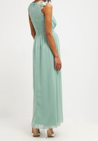 Maxi-mekko vaaleanvihreästä sifongista, jossa on pitsiyksityiskohdat olkapäillä, tiukka yläosa ja valuva helma, ylös kerätty vyötärödetalji.