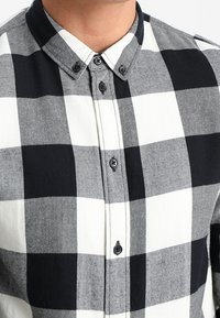 Camisa de cuadros en blanco y negro con cuello puntiagudo, que presenta grandes patrones de cuadros, botones negros y una textura de algodón suave.