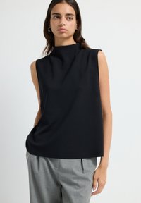 Jeune femme avec des cheveux bruns lisses, portant un haut sans manches noir et un pantalon plissé gris, sur fond blanc uni.