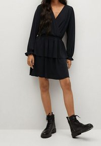 Vestido negro de manga larga con cintura peplum y textura suave, combinado con botas de tobillo negras con cordones y suela gruesa.