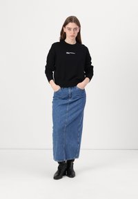Črna sweatshirt z žametenimi manšetami in okroglo izrezano ovratnikom, ki ima belo logotipsko besedilo. V kombinaciji z dolgo modro denim krilom in črnimi gležnjarji.