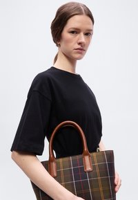 Γυναίκα με ανοιχτόχρωμο δέρμα και καστανά μαλλιά, με μαύρο πουκάμισο, κρατώντας μια καρό τσάντα tote με καφέ δερμάτινη λαβή, μπροστά σε απλό φωτεινό φόντο.