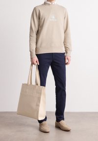 Persona che indossa un maglione beige con logo Calvin Klein, pantaloni blu navy, mocassini beige, mentre tiene una grande borsa tote beige, in piedi su un pavimento chiaro.