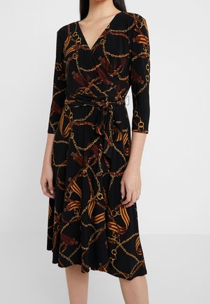 Kobieta w czarnej sukience kopertowej z nadrukiem brązowych i złotych łańcuchów oraz paska, z rękawami do połowy przedramienia i zawiązywanym paskiem w talii.