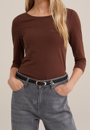 Kobieta ubrana w brązową bluzkę z długim rękawem wpuszczoną do szarych dżinsów z czarnym paskiem, ręce spoczywające w przednich kieszeniach.