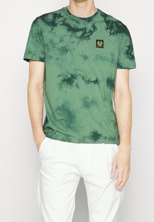Homme portant une chemise tie-dye verte à manches courtes avec un patch logo sur la poitrine et un pantalon blanc, debout les mains dans les poches.