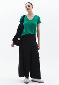 Camiseta verde de cuello en V combinada con pantalones negros holgados, acentuada por un bolso de mano negro. Zapatillas blancas completan el atuendo. Diseño minimalista.