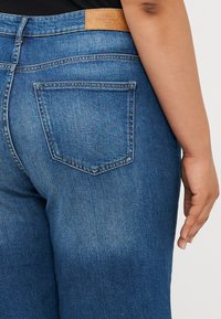 Sötétkék denim farmernadrág egyenes szabással, derékrészen bőr címkével és két hátsó zsebbel, egyszerű varrásmintával.