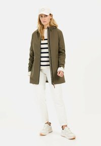 Olive green waterproof jacket with a front zipper and hood, worn over a white and navy striped sweater, paired with white pants and sneakers.