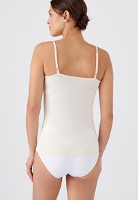 Canotta beige con spalline sottili, realizzata in un tessuto liscio. Presenta una linea dritta e una texture sottile. Abbinata a un pantalone bianco.