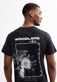 Camiseta de algodón negra con un diseño gráfico blanco de flores y el texto "WOODLAND vibes" en la parte posterior. Cuello redondo clásico y mangas cortas.