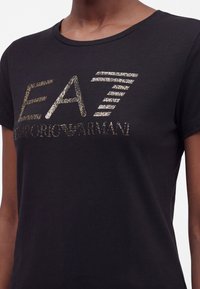 Άτομο που φοράει μαύρο T-shirt με μεταλλικό χρυσό λογότυπο "EA7 Emporio Armani" τυπωμένο στο στήθος.