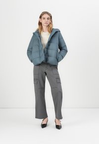 Padded jacket v tlumené modré barvě, se zipem a kapucí. Nosi se s šedými cargo kalhotami s kapsami a černými špičatými podpatky.