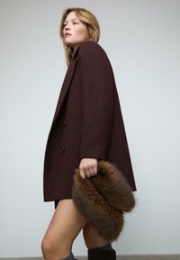 Donna che indossa un blazer marrone scuro e calzini grigi, che tiene un grande accessorio di pelliccia marrone contro uno sfondo chiaro e semplice.