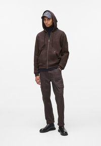 Rjava zip-up kapuca z kengurujskim žepom, v kombinaciji z usklajenimi cargo hlačami z več žepi. Dokončano z črnimi čevlji in sivo kapo.