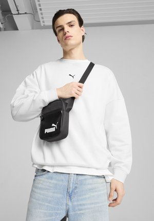 Bolso negro de bandolera con el logo de PUMA, hecho de material sintético, llevado sobre una sudadera blanca de corte relajado, combinado con jeans azul claro.