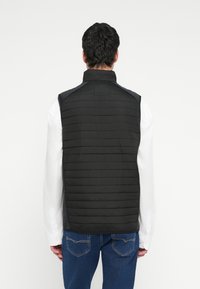 Čierna prešívaná vesta s vertikálnym prešívaným vzorom, vysokým golierom a bez rukávov, nosená cez bielu košeľu s dlhým rukávom, v kombinácii s modrými džínsami.