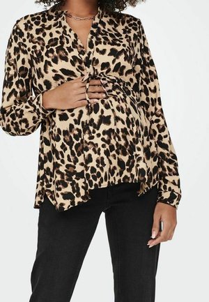 Kvinde iført leopardmønstret ventebluse bundet over maven med lange ærmer og sorte bukser.