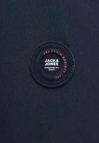 Crni materijal s okruglom etiketom koja sadrži bijeli i crveni tekst: "JACK & JONES," "J&J DENIM SOCIETY," i broj zaštitnog znaka.