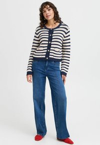 Pruhovaný cardigan v námořnické modři a krémové barvě s kulatým výstřihem, předními knoflíky a kapsami. V kombinaci s rozšířenými modrými džíny a červenými botami.