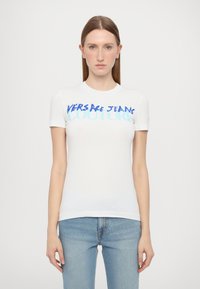Bijela pamučna majica kratkih rukava s podebljanim plavim natpisom "VERSACE JEANS COUTURE" preko prsiju. Prilagođen kroj s glatkom teksturom.