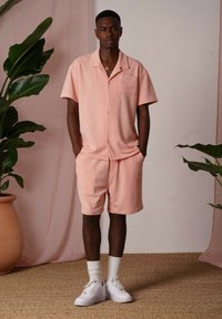 Camicia button-up rosa pallido a maniche corte con tasca, abbinata a pantaloni corti coordinati. Indossata con sneakers bianche e calze alla caviglia.