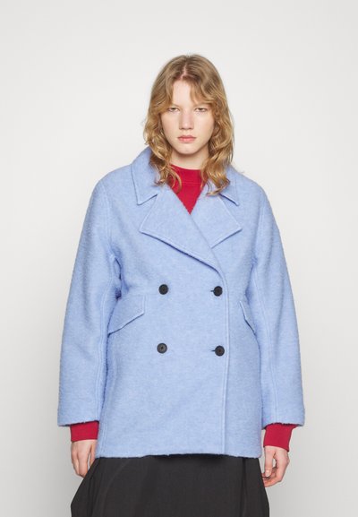 Jeune femme aux cheveux blonds ondulés portant un manteau en laine bleu à double boutonnage, un pull rouge en dessous et une jupe noire, se tenant devant un fond uni.