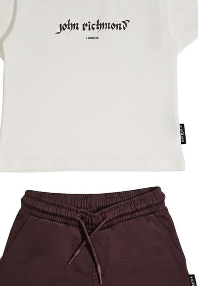 T-shirt bianca corta con la scritta "john richmond LONDON" sopra abbinata a pantaloncini con coulisse e fascia elastica in vita di colore marrone scuro.