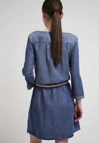 Nainen, jolla on pitkät ruskeat hiukset poninhännällä, pukeutuu siniseen farkkuhamoseen, jonka hihat on kääritty ylös ja vyöllä on letitetty vyö, selin yksinkertaista taustaa vasten.