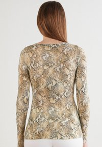 Vrouw met lang bruin haar, gekleed in een strakke, lange mouwen, slangenprint top, gezien van de achterkant tegen een effen achtergrond.