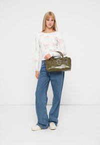 Olivgrüne Handtasche mit einem sternförmigen Akzent, glatter Lederstruktur und stabilen Griffen. Das Model trägt ein bedrucktes Langarmoberteil und Jeans.