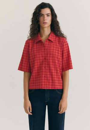 Frau mit langen lockigen Haaren, die ein rotes kurzärmeliges Hemd mit Knöpfen und dunkle Jeans trägt, steht vor einem schlichten hellen Hintergrund.