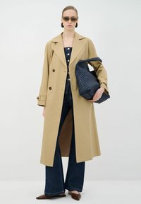 Tan dubbelzijdig trenchcoat, donkerblauwe denim flared broek, geweven marineblauwe tas en bruine puntige schoenen; vergezeld van een zonnebril.