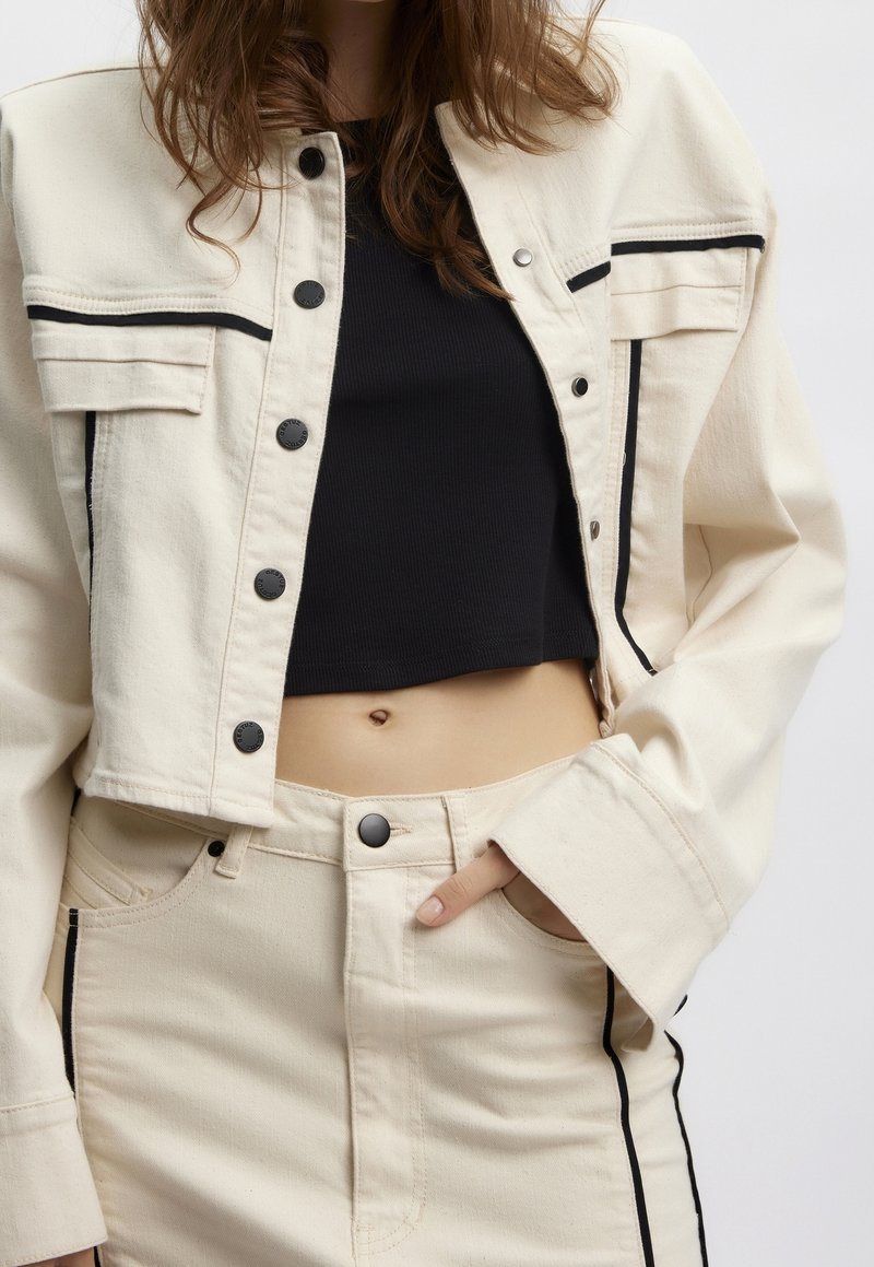 Kobieta ubrana w kremową kurtkę i spodnie z denimu z czarną lamówką, czarny krótki top, ręka w kieszeni spodni, na tle jednolitego białego tła.