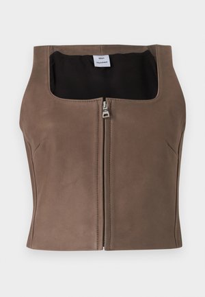 Ženska z rjavim brezrokavnikom z zadrgo spredaj, širokimi modrimi kavbojkami s črnim pasom in črnimi zaobljenimi čevlji, stoji z rokami v žepih.