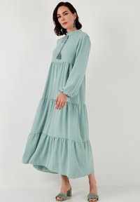 A long, mint green dress with long sleeves, tiered design, a tassel detail at the neckline, and soft, flowing fabric.