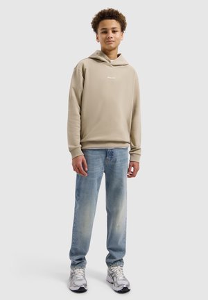 Tienerjongen staand, gekleed in een beige capuchontrui, lichtblauwe spijkerbroek en witte sneakers tegen een effen grijze achtergrond.