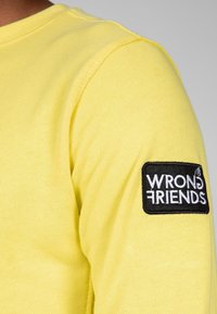 Camisa amarilla de manga larga con textura acanalada, que presenta un parche negro con el texto blanco "amigos equivocados" en el hombro izquierdo.