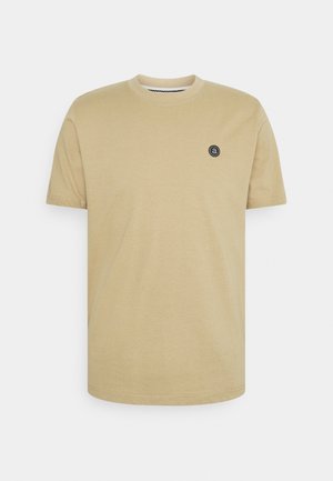 T-shirt beige à manches courtes et col rond avec un petit logo noir circulaire sur le côté gauche de la poitrine, sur fond uni.
