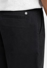 Pantalon noir en tissu lisse, avec une poche arrière portant une étiquette logo et une poche latérale. Le design est épuré et simple.