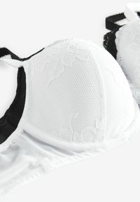 Soutien-gorge blanc et noir avec détails en dentelle et superposition en tulle. Il présente des bonnets incurvés et des bretelles réglables, mettant en valeur une surface texturée.