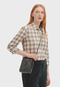Giovane donna con lunghi capelli rossi, che indossa una camicia a quadri beige e pantaloni scuri, mentre tiene una piccola borsa a tracolla nera sulla spalla.